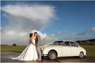 bride and groom kissing next to vintage jaguar car against dramatic blue sky